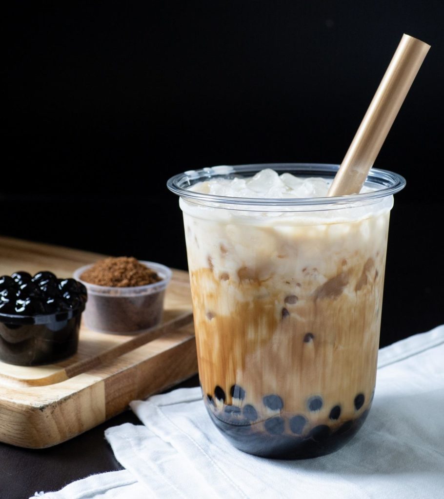 Satu gelas bubble tea dengan boba dan brown sugar di belakangnya.