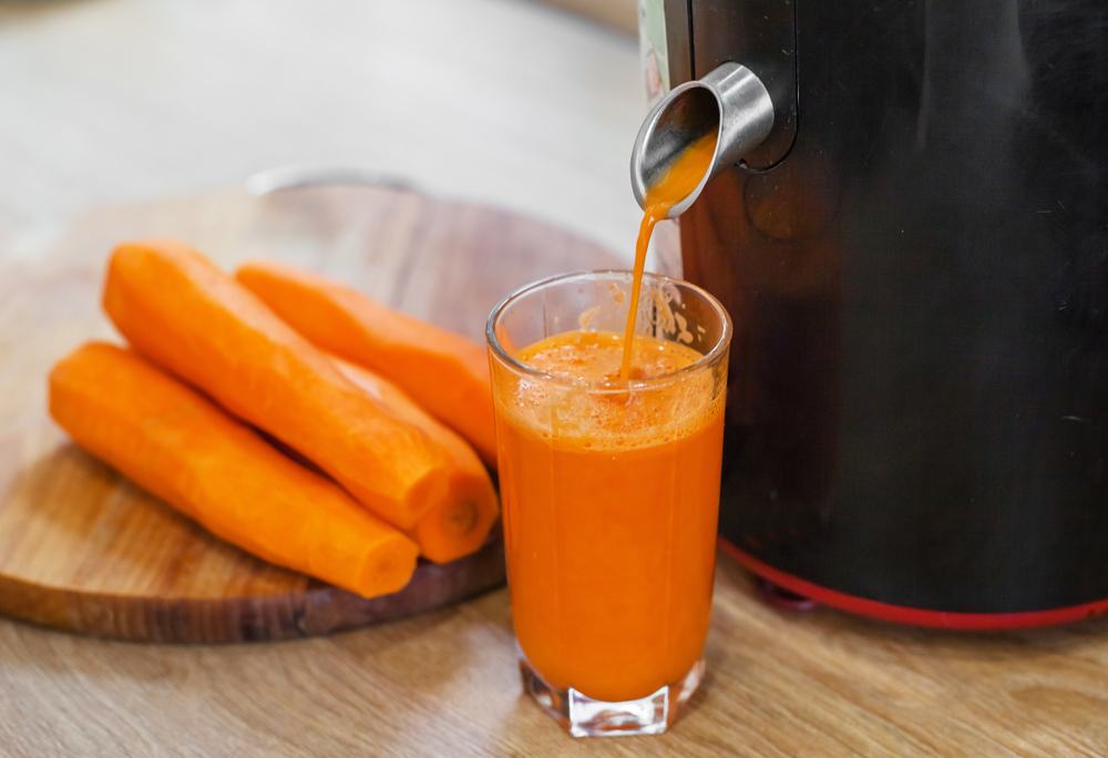 Pressed juicer mengeluarkan jus wortel ke dalam gelas bening dengan wortel utuh di belakangnya.