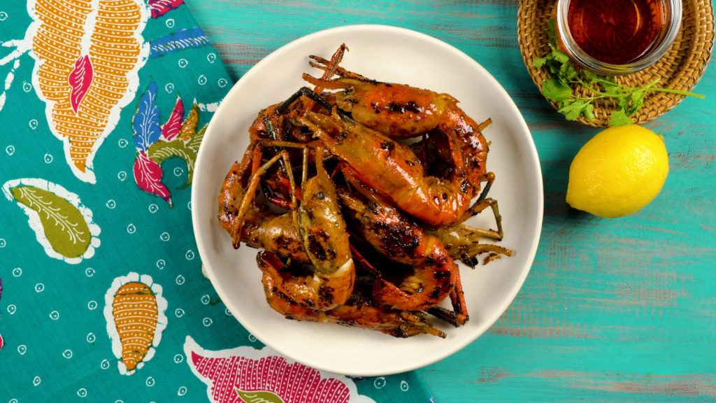 Udang bakar madu tersaji di atas piring dan latar bahan serta kain batik.