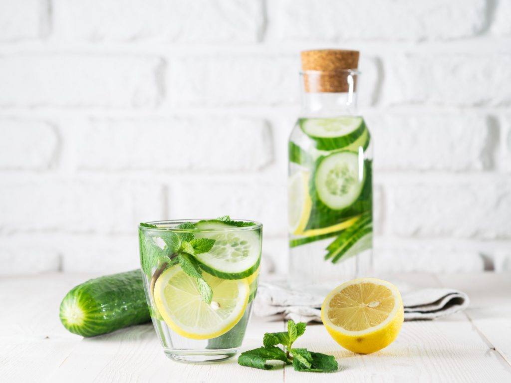 Resep Infused water untuk detox berupa lemon, mentimun, dan daun mint dalam gelas mening, dengan latas botol bening berisi infused water yang sama.