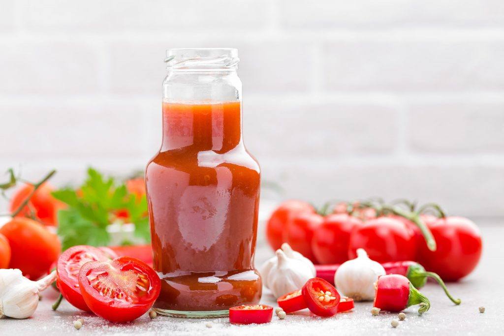 Botol kaca berisi saus tomat dengan potongan tomat di sektiarnya.