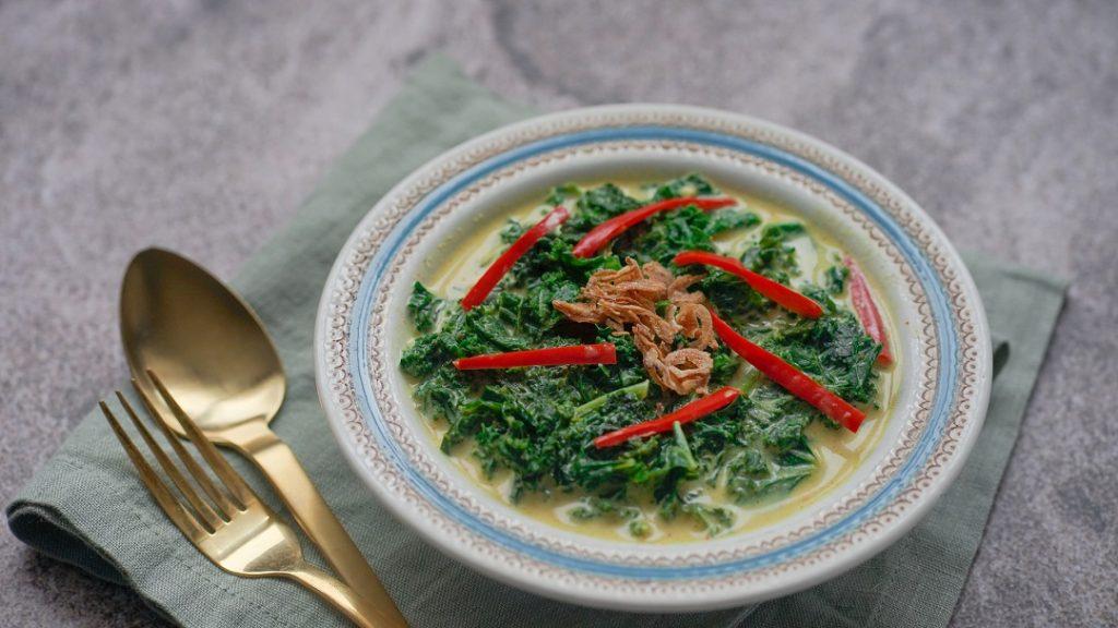 Seporsi gulai daun singkong tersaji dalam mangkuk dan didampingi alat makan emas.