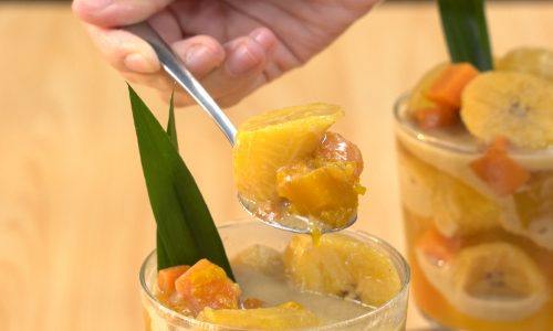 Kolak pisang milk tea tersaji di dalam gelas yang berhiaskan daun pandan dan tengah dinikmati menggunakan sendok.