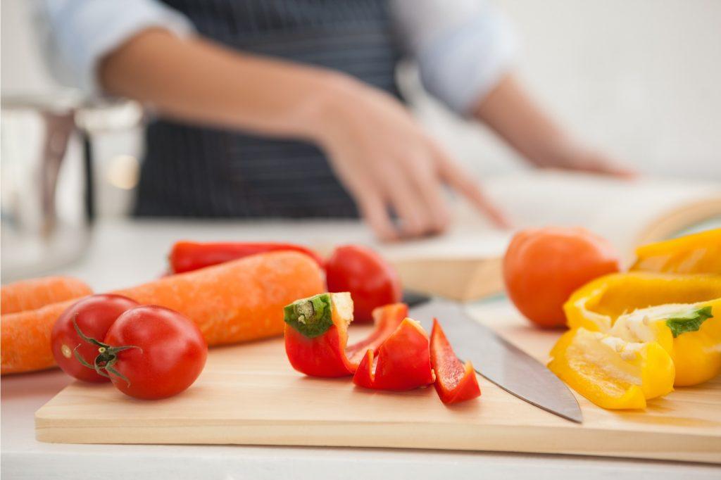 Tangan wanita sedang menunjuk buku resep di atas meja dapur dan berserakan juga sayuran yang akan dipotong.