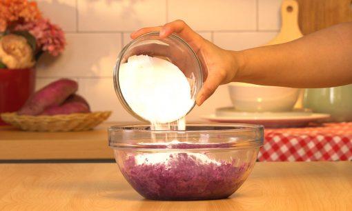Tepung dituangkan ke dalam ubi ungu yang telah dihaluskan dalam mangkuk kaca.