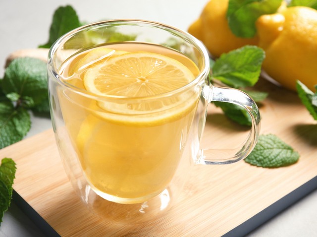 Segelas air lemon hangat tersaji dia atas nampan kayu dan didampingi buah lemon serta daun mint.
