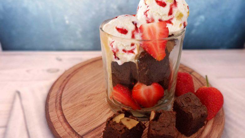 Satu gelas berisi Strawberry Cheesecake Brownie Sundae yang tersaji bersama buah stroberi segar.