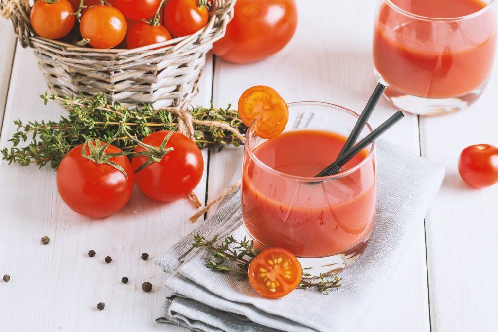 Menikmati manfaat jus tomat dalam gelas bening dengan tomat segar di belakangnya.