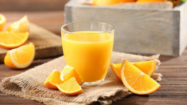 Segelas jeruk hangat segar didampingi potongan buah dan disajikan di atas napkin serta meja kayu.