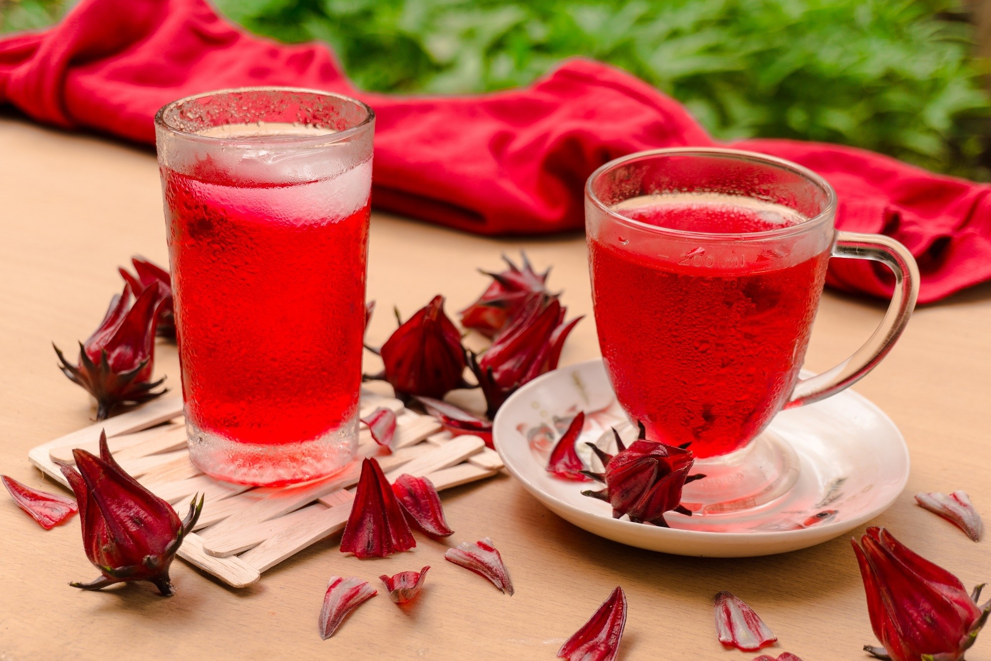 Dua gelas kaca berisi hasil membuat resep es teh rosella dengan bunga rosella kering di bawahnya