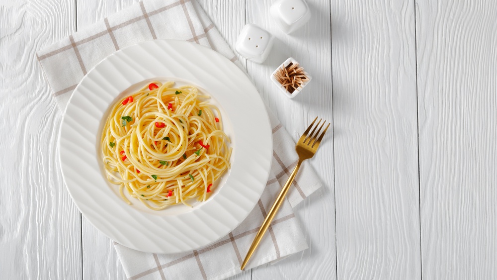 Resep aglio olio disajikan dalam piring putih di atas serbet dan meja kayu putih.