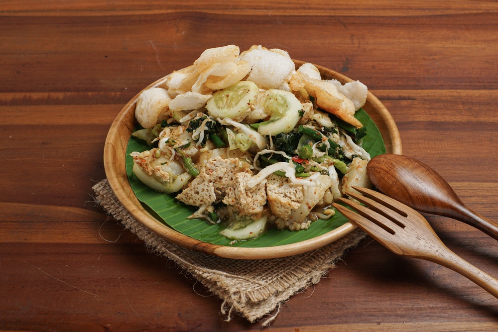 Hasil masak menu sayur resep lotek Bandung beralas daun pisang hijau di piring cokelat dan sendok garpu.