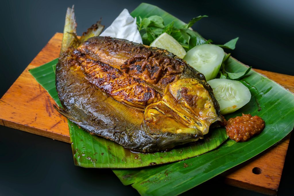 Ikan bakar di atas daun pisang, dilengkapi potongan timur dan sambal.