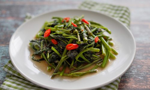 Tumis kangkung terasi dalam piring saji putih dialasi serbet warna hijau putih.